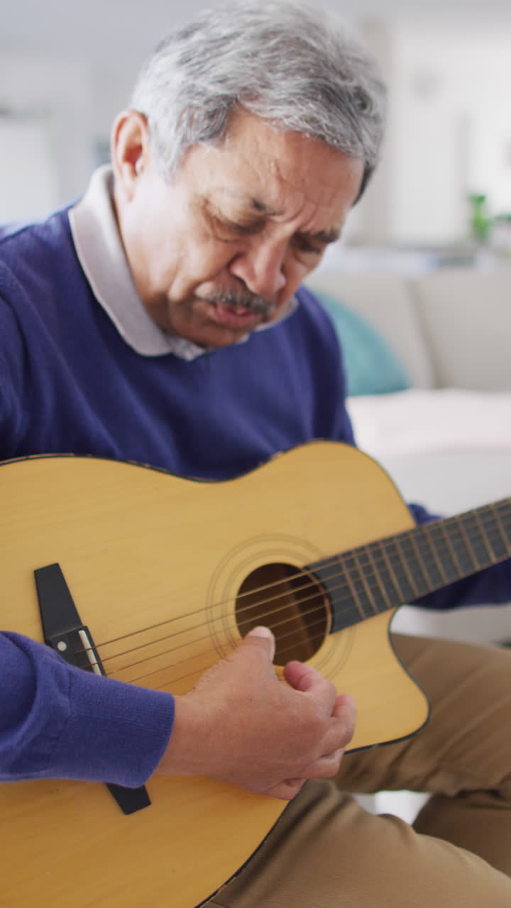 video vertical de un anciano biracial cantando y tocando la guitarra acústica, sentado en casa