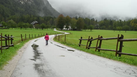 Kleines-Mädchen,-Das-Im-Regen-Auf-Einer-Landstraße-Läuft,-Bauernhaus-Im-Hintergrund,-Alpental,-4K-Aufnahme,-Von-Hinten-Weggewandt