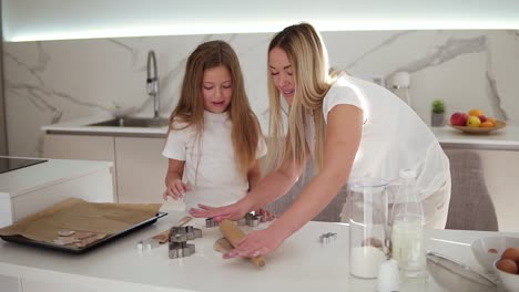 Caucasian-good-looking-mother---blonde-hair,-wearing-white-T-shirt-teaching-her-daughter-to-roll-a-dough-in-the-cozy-modern-white-kitchen.-Girl-cutting-a-shaped-cookies.-Slow-motion
