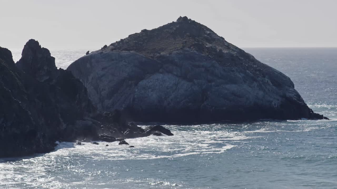 Ocean waves swirl around a rugged sea stack off California’s Big Sur coast, as light glints off the dark rock and surrounding sea spray adds atmosphere.