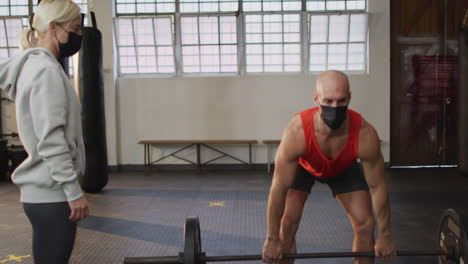 caucasian muscular man wearing face mask and lifting weight bar with coach