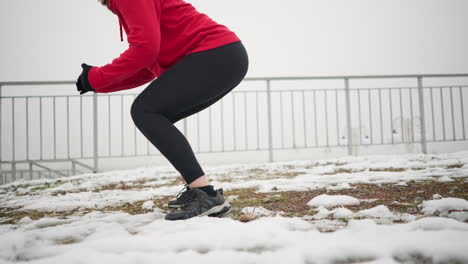 woman performing jump squats on snowy ground, wearing red hoodie and black gloves, focusing on leg strength, background includes snowy environment with metal railing and foggy atmosphere