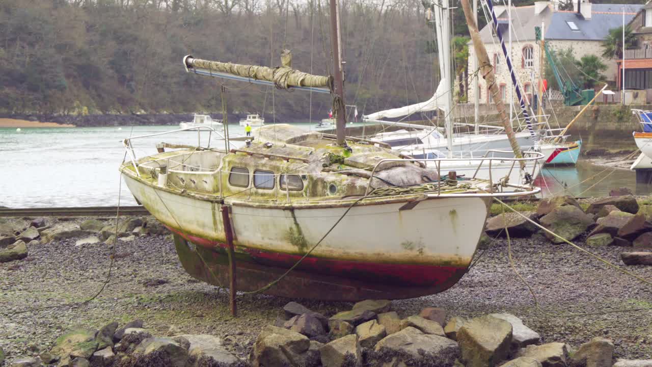 A weathered sailboat with moss-covered hull and peeling paint rests on a rocky shore at low tide. The winter setting feels cold and still, evoking abandonment and time’s passage.