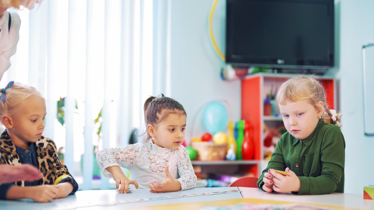 Little girls during the lesson on the classroom background with TV. Primary students playing picture cards game with the help of the teacher in class.
