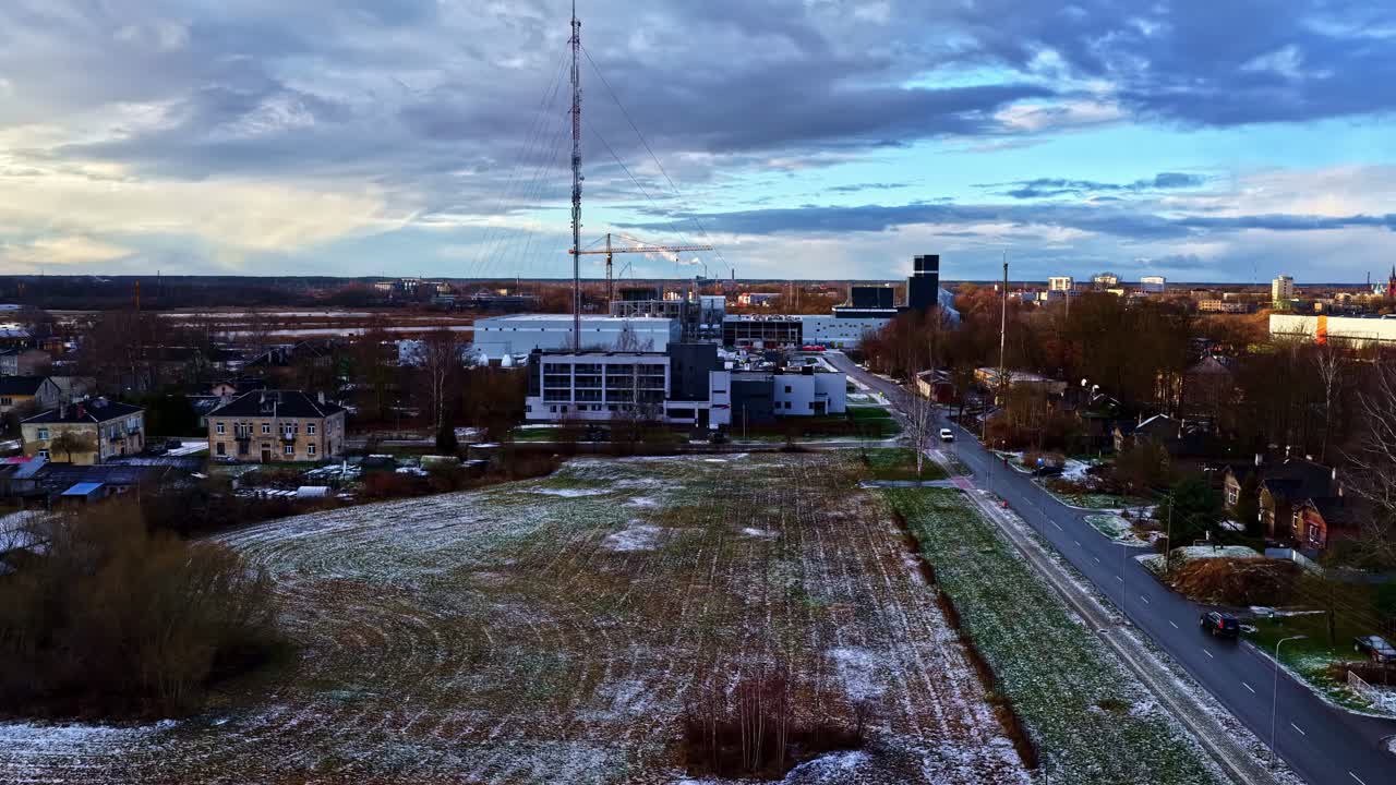 Sorvolo di una zona industriale periferica con drone, con torre radio, fabbriche e strada suburbana accanto a un campo gelido sotto nuvole invernali suggestive durante l'ora d'oro