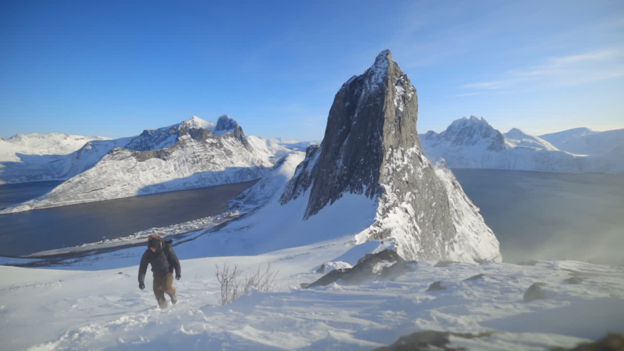 un hombre escalando una empinada ladera nevada con fuertes vientos que soplan lentamente y el monte segla en el fondo, cámara lenta
