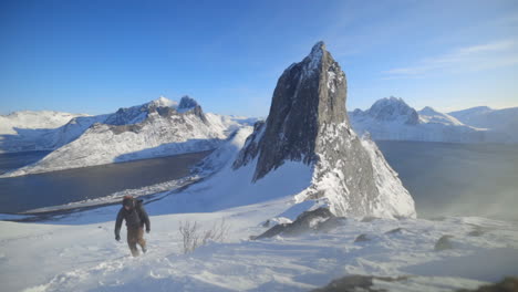 a man hiking up a steep snowy mountainside with high winds blowing slow and mount segla in the background, slow motion