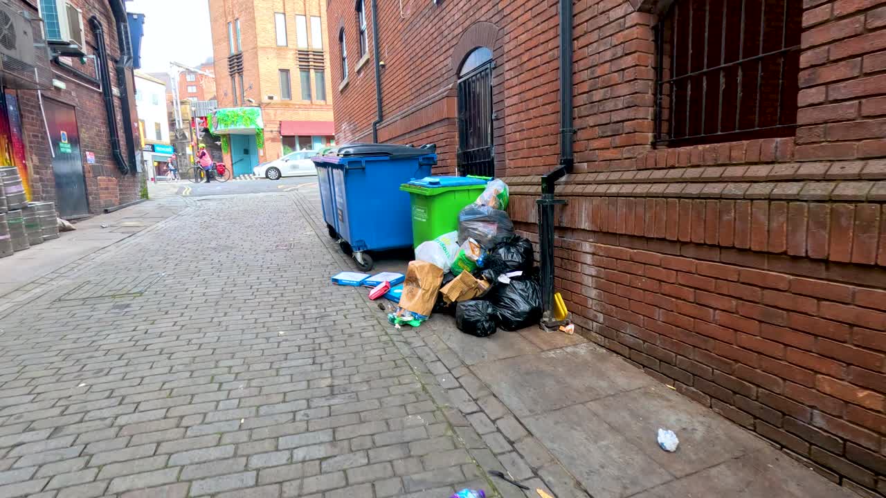 Overflowing blue and green rubbish bins line a cobblestone alley beside brick buildings, with scattered trash and colorful graffiti under natural daylight. Camera slowly moves forward