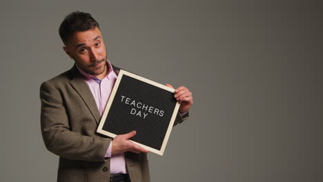 studio portrait of mature male teacher standing against grey background holding up notice board reading teachers day