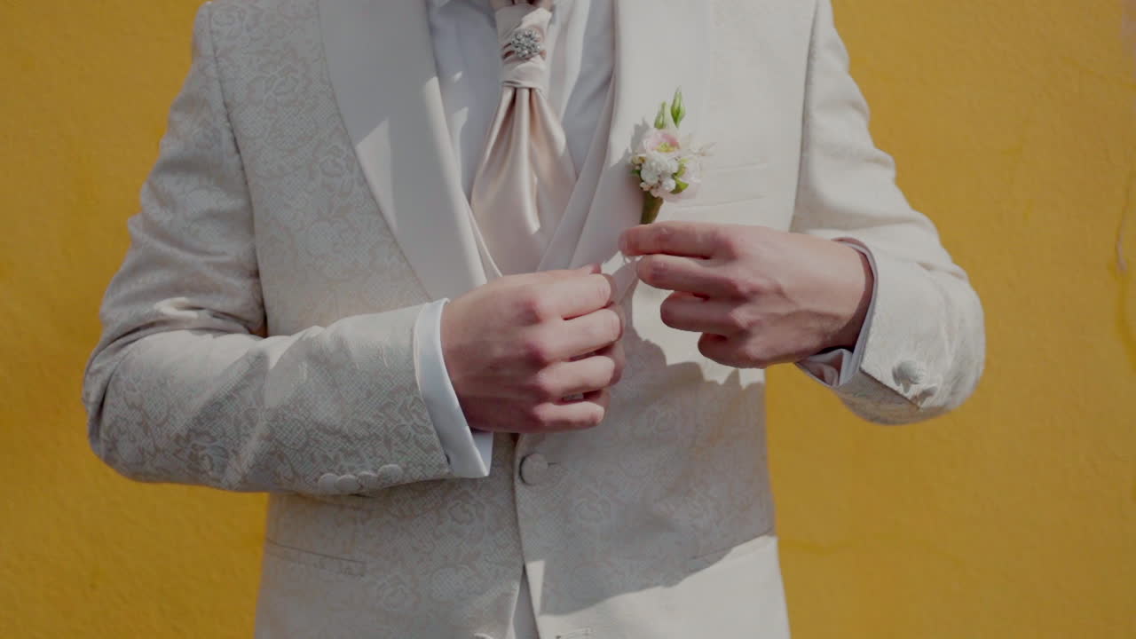 색 꽃복을 입은 신랑이 활기찬 노란색 배경을 배경으로 부토니에를 조정하고 있습니다.