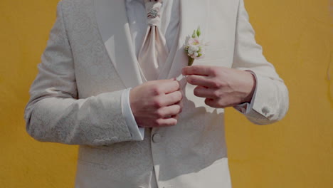 groom in white floral suit adjusting boutonniere, set against a vibrant yellow background