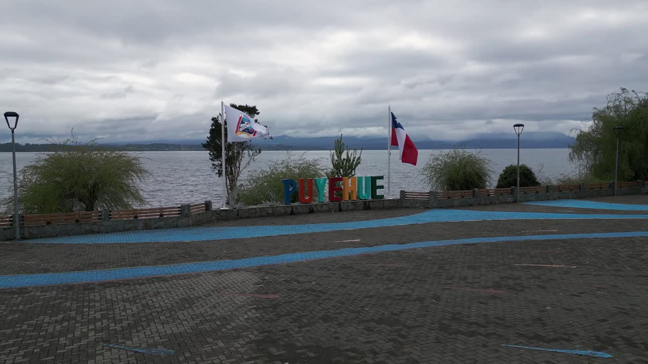Toma aérea en órbita de una plaza frente al lago con coloridas letras de Puyehue y banderas chilenas junto a estructuras de sombra azules con vistas a las tranquilas aguas del lago bajo un cielo nublado dramático. Entre Lagos, Puyehue, Chile