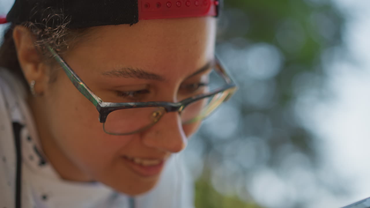 retrato enfocado de una mujer jugando; las gafas reflejan la pantalla; sonrisa sutil; cable del auricular visible; fotos de perfil concentradas en un parque; pensamiento táctico; práctica de niveles; bokeh a la luz del día; natural
