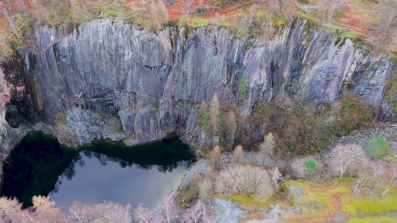A drone gently descends toward a quarry lake, shifting focus from treetops and winter colors above to steep slate walls and the still water below in a dramatic downward reveal