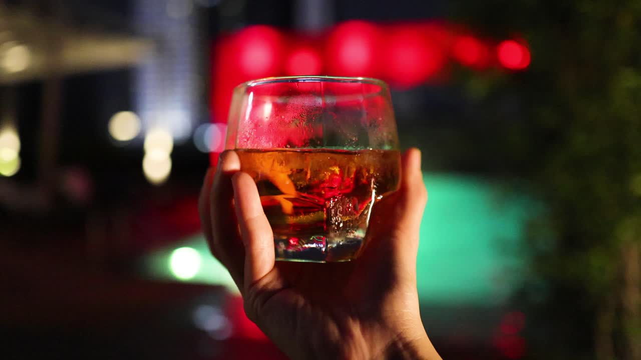 A hand slowly rotates a cocktail glass with ice under colorful, blurred nightlife lights in an outdoor Bangkok bar. Warm, moody lighting and shallow depth of field