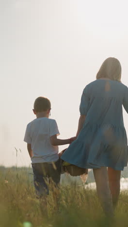a back shot of a mother in a blue dress carrying a basket in one hand holding her younger son's hand with the other. her older son walks beside them as they walk through a grassy field towards a lake