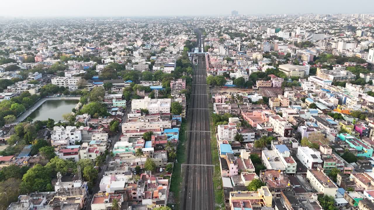 Aerial video of Chennai, India, showing vast urban sprawl. A long stretch of railway tracks cuts through the dense cityscape, with a serene temple tank and lush greenery providing a peaceful contrast