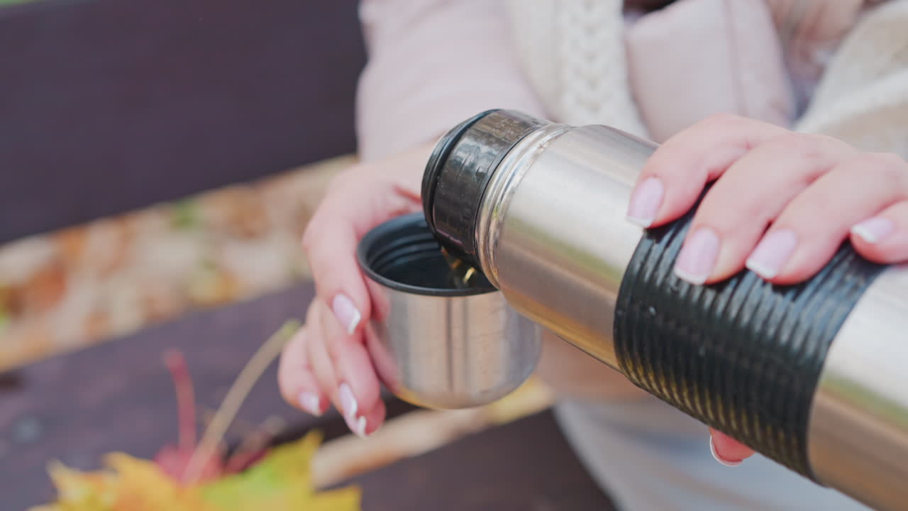 Close up of woman pouring hot beverage from thermos into small cup while sitting on bench in autumn park. Focus on hands, thermos, cup, and cozy clothing with fallen leaves in soft background blur