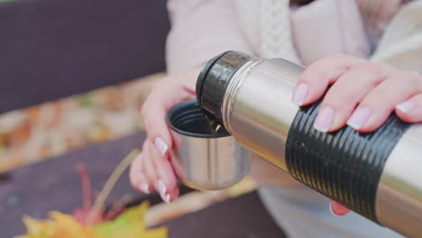 close up of woman pouring hot beverage from thermos into small cup while sitting on bench in autumn park. focus on hands, thermos, cup, and cozy clothing with fallen leaves in soft background blur