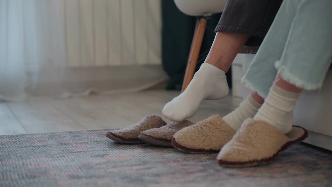 Piedi del tutor accanto alle pantofole. Studente e tutor sono seduti su sedie, su un tappeto morbido, con pantofole pelose e jeans di denim, in una calma sessione di coaching di grammatica a casa. Guida paziente, luce naturale calda, una scena di studio domestica intima.