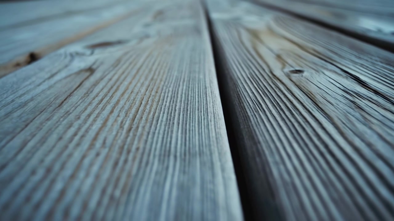 Close-up of gray weathered deck boards running diagonally across the frame, showing rough wood grain, seams, and natural aging with a softly blurred background