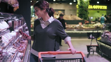 young brunette female costumer looking at cellphone while walking through meat aisle in grocery store. girl takes salami stick from the shelf and puts it into shopping cart