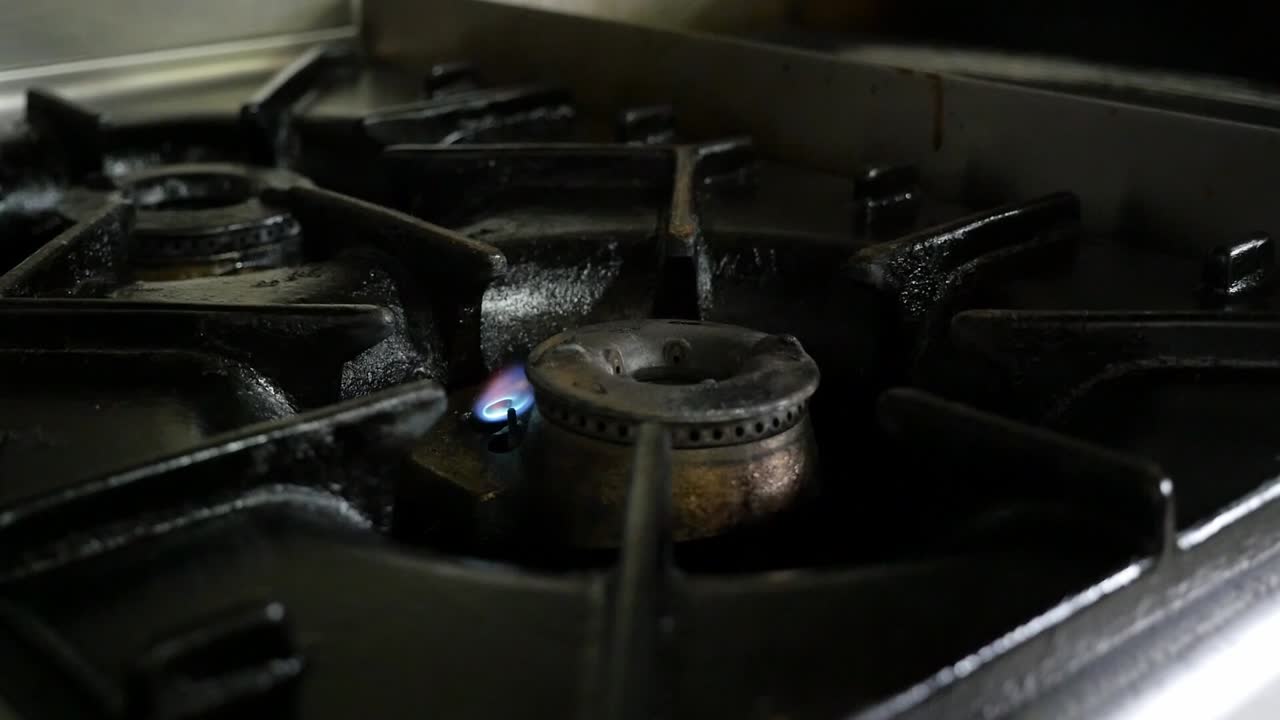 Close-up of an industrial kitchen burner being lit. A stainless steel pan with salmon is placed on the burner to begin cooking