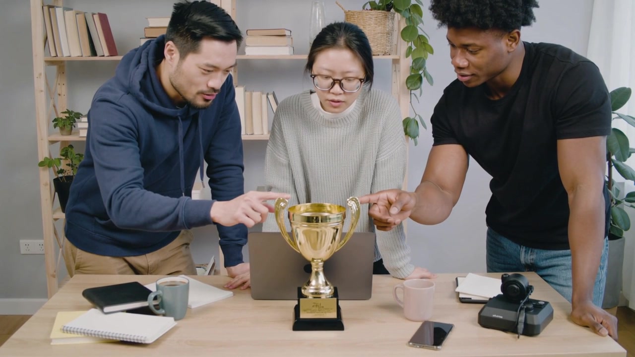 Reaching teammate in hoodie adjusting gold trophy at office, center checking laptop confirming win