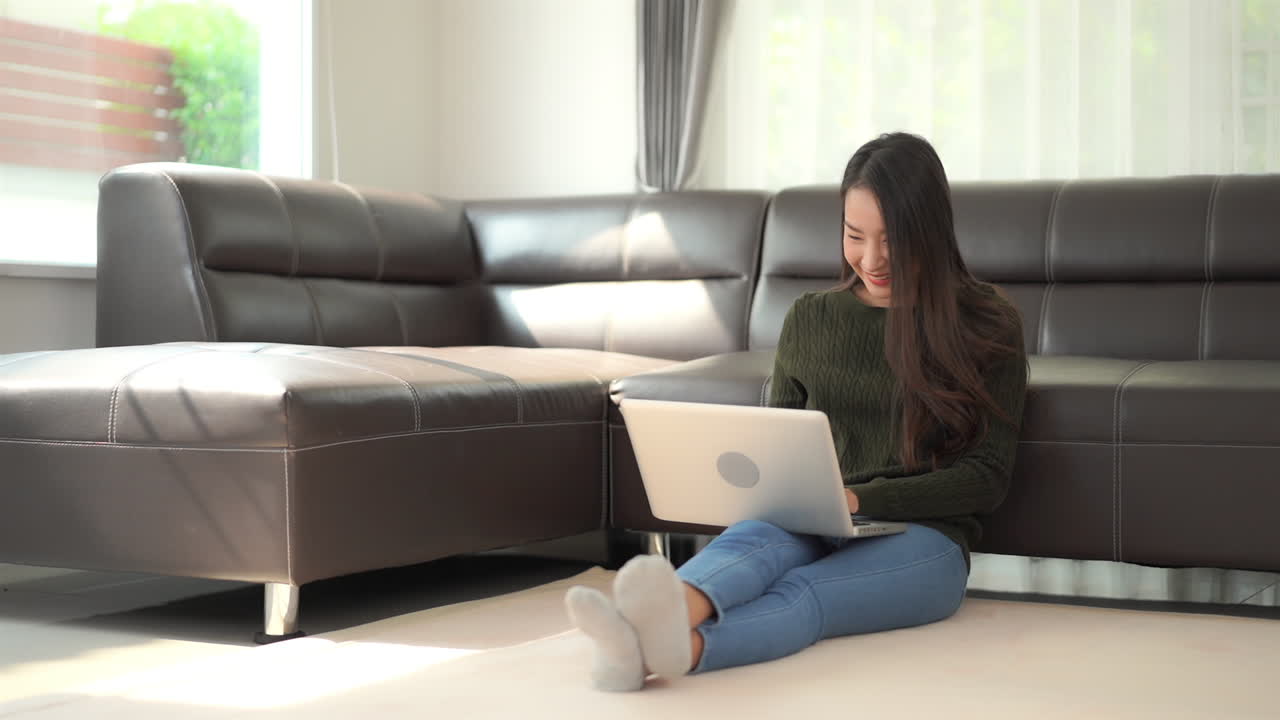 Happy woman pumps fist showing success while sitting on living room floor with laptop computer. Asian model in casual clothes sits on floor against sofa, smiles and celebrates win or good news