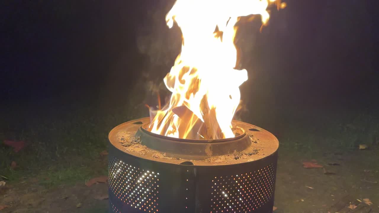 Large flames bursting from circular metal fire pit in dark outdoor setting