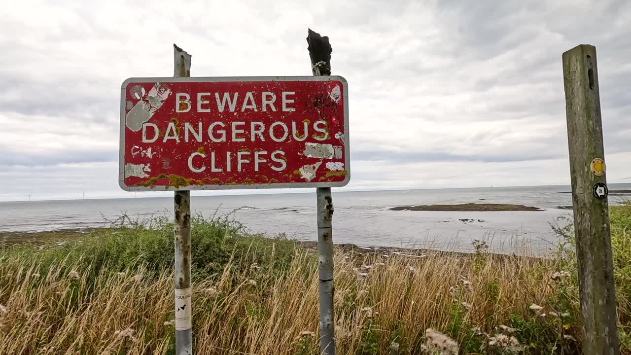 A red warning sign reading 'Beware Dangerous Cliffs' stands on a grassy, rocky shoreline under cloudy daylight, with slow camera movement approaching the sign