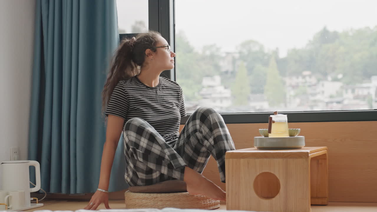Donna alla finestra dell'hotel che si gira verso la vista, ospite studente che esplora il davanzale e si siede a contemplare il panorama della città, pantaloni casual a quadri, luce che filtra dalle tende, piccolo vassoio di legno sul tavolino laterale.
