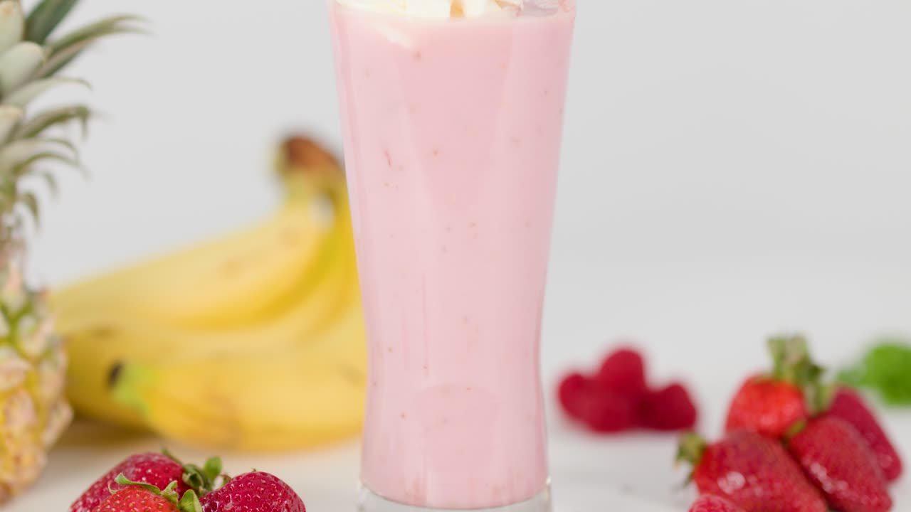 Whipped cream added to pink smoothie in tall glass, surrounded by fresh fruit, bright lighting