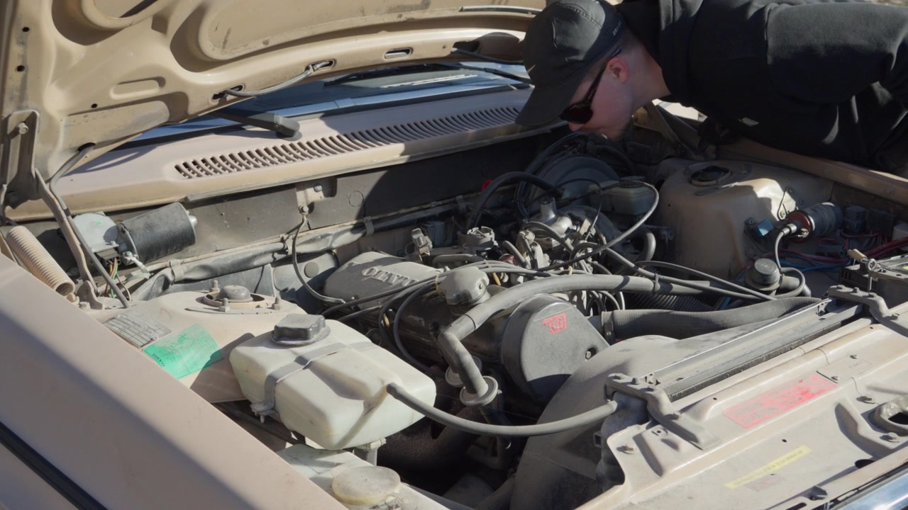 A mechanic inspects an old carbureted engine under the open hood, showing hands-on maintenance of an aging gasoline vehicle.