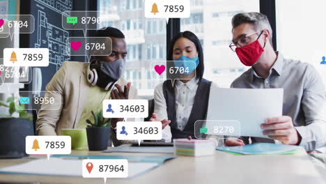 Animation-of-social-media-icons-over-diverse-colleagues-wearing-face-masks-discussing-at-office