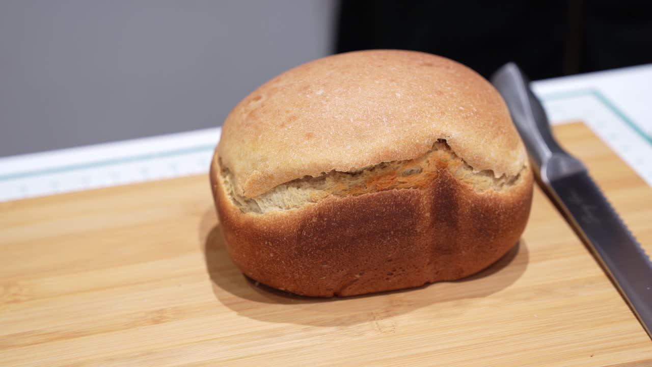 Close up of a freshly baked homemade bread loaf with golden crust placed on a wooden cutting board beside a kitchen knife, highlighting simple baking and food preparation concept