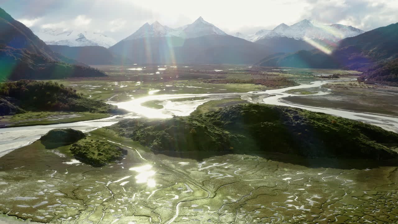 Sunlight reflects off wet Patagonian terrain in broad valley near river junction at golden hour, glistening wetland meanders below ridge