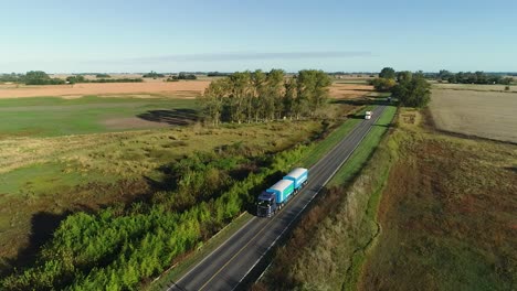 Toma-Aérea-De-Camiones-Circulando-Por-Una-Carretera-Rural-En-Un-Día-Soleado,-Proyectando-Largas-Sombras