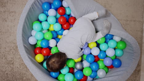 adorable toddler child falls on the pool filled with colorful balls. kid dives into balls moving in them with smile. top view.