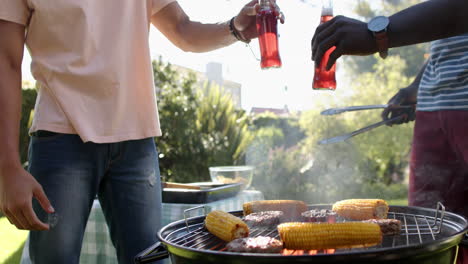 grilling corn and meat, two multiracial male friends toasting with drinks at outdoor barbecue