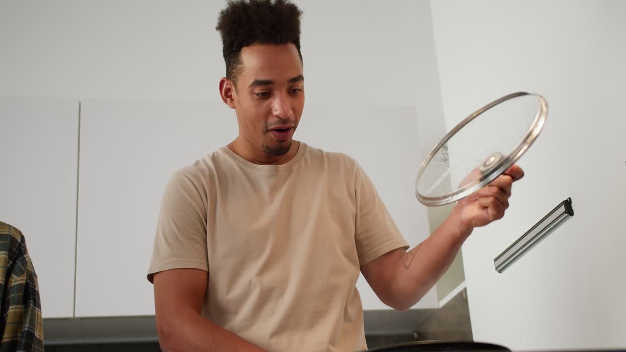 A happy young man with Black skin in a beige T-shirt lifts the lid of a frying pan and a large amount of steam comes out from there while preparing breakfast with his young adult brunette girlfriend with a bob hairstyle in a modern kitchen