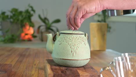 hacer té de hierbas en una tetera verde sobre una mesa de madera en una habitación luminosa y aireada con plantas verdes en el fondo y una taza de té de vidrio