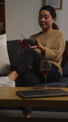 vertical video of woman at home sitting on sofa eating healthy vegetarian or vegan meal of tabbouleh salad with glass of wine with mobile phone and laptop on table