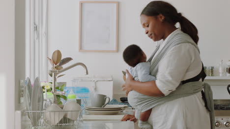 african american mother holding baby working at home washing dishes cleaning kitchen caring for toddler doing housework enjoying motherhood