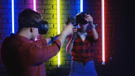 young man and woman in vr glasses and using joystick while playing a virtual reality game in a room with colorful neon lamps on the wall 2