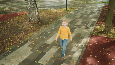 young boy wearing orange shirt and jeans walking alone through quiet park, looking around curiously while crossing wet pavement scattered with autumn leaves and surrounded by trees