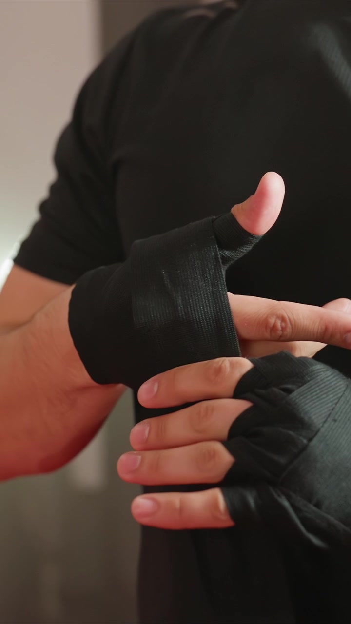 Primo piano di un uomo che si fascia le mani con bende nere. Il processo prevede una fasciatura dettagliata. Ambiente interno con illuminazione soffusa e sfondo neutro che enfatizza i dettagli in primo piano.