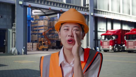 close up of asian female engineer with safety helmet yelling with hand over mouth, outside of logistics distributions warehouse