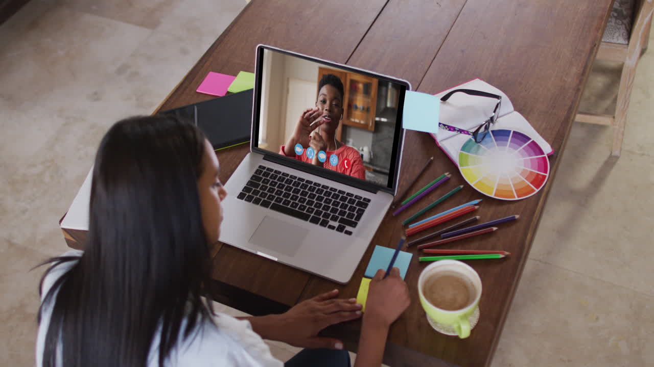 Mujer caucásica usando una laptop en videollamada con una colega y tomando notas
