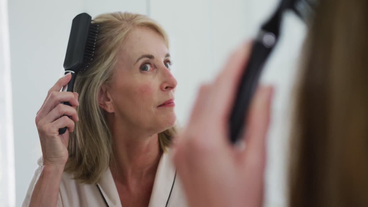 Mujer mayor caucásica mirándose en el espejo del baño y peinándose el cabello.